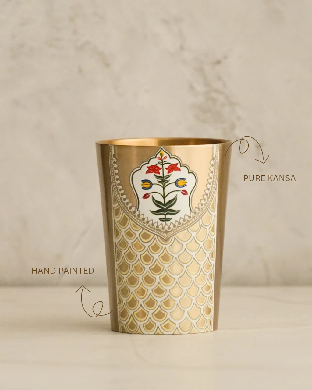 A gold-toned metal cup with hand-painted floral and fish-scale patterns, shown on a light marble surface with text labels pointing to its finish.