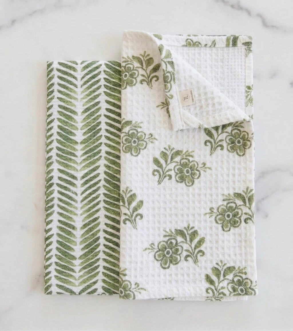 A flat-lay view of two white waffle-weave towels with green botanical prints. One towel has a floral motif and a folded corner, while the other features a vertical leaf pattern, both set against a light marble background.