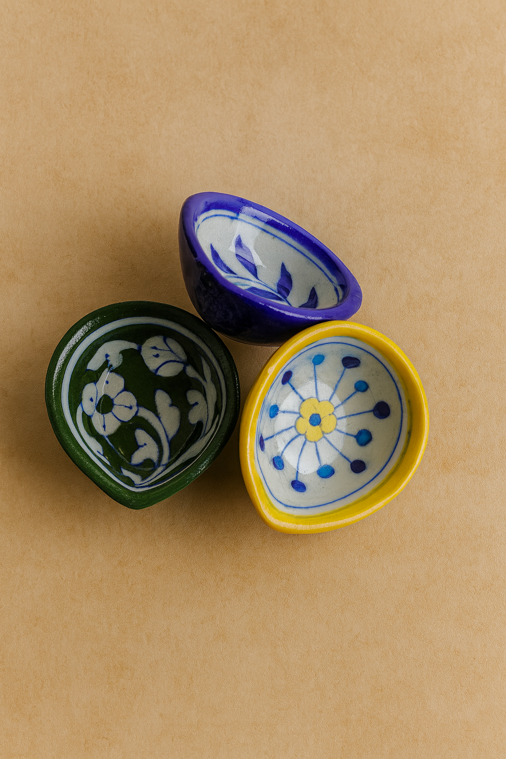 Three ceramic diya (yellow, blue, green) with floral patterns on a beige background